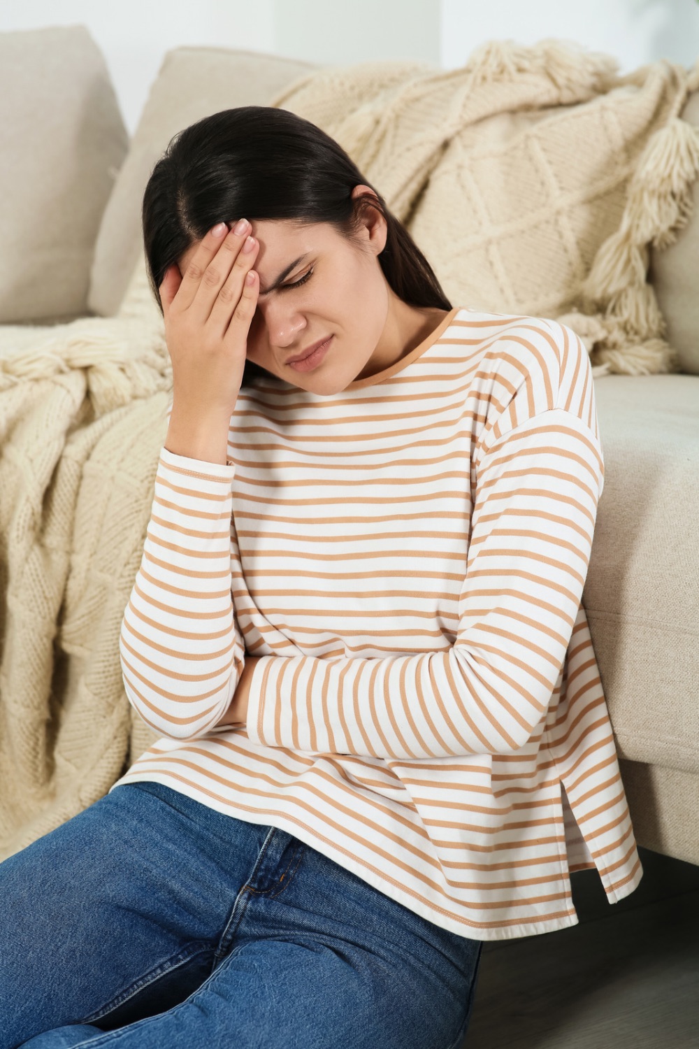 Woman wearing a stripy top and jeans sitting on floor in front of the sofa holding her forehead and belly