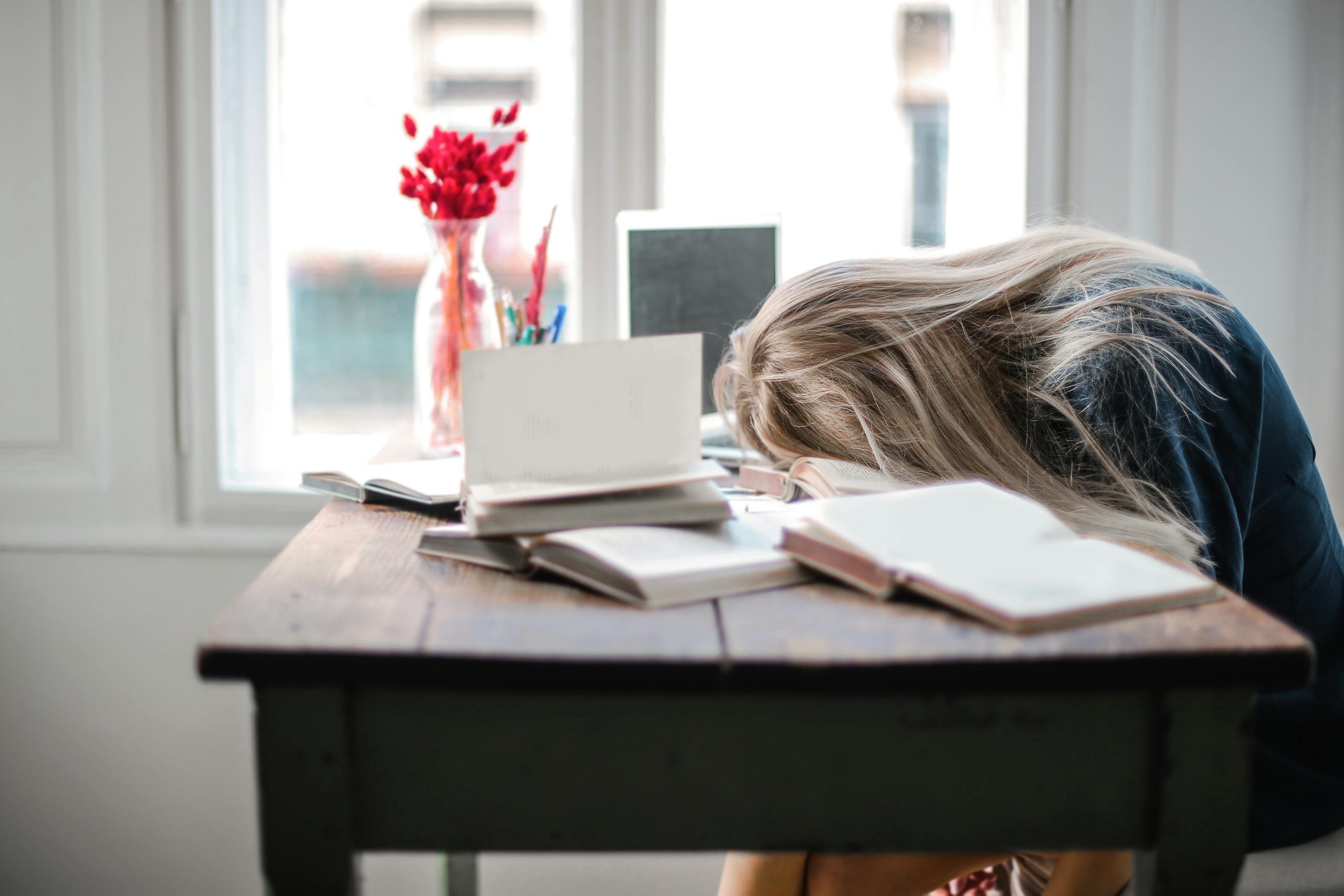 Blonde woman slumped over desk filled with books, showcasing study fatigue indoors, with poor metabolic health and energy levels.