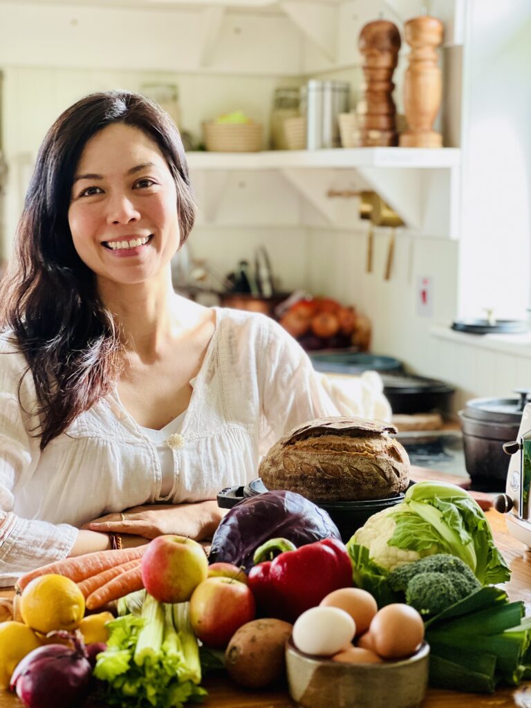 woman standing in cottage kitchen wearing white blouse, with nutritious food like fruit, vegetables and eggs in the foreground and open shelving in the background.