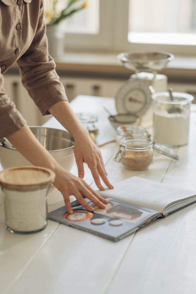 A woman in a cozy kitchen flipping through a cookbook while gathering ingredients for baking.