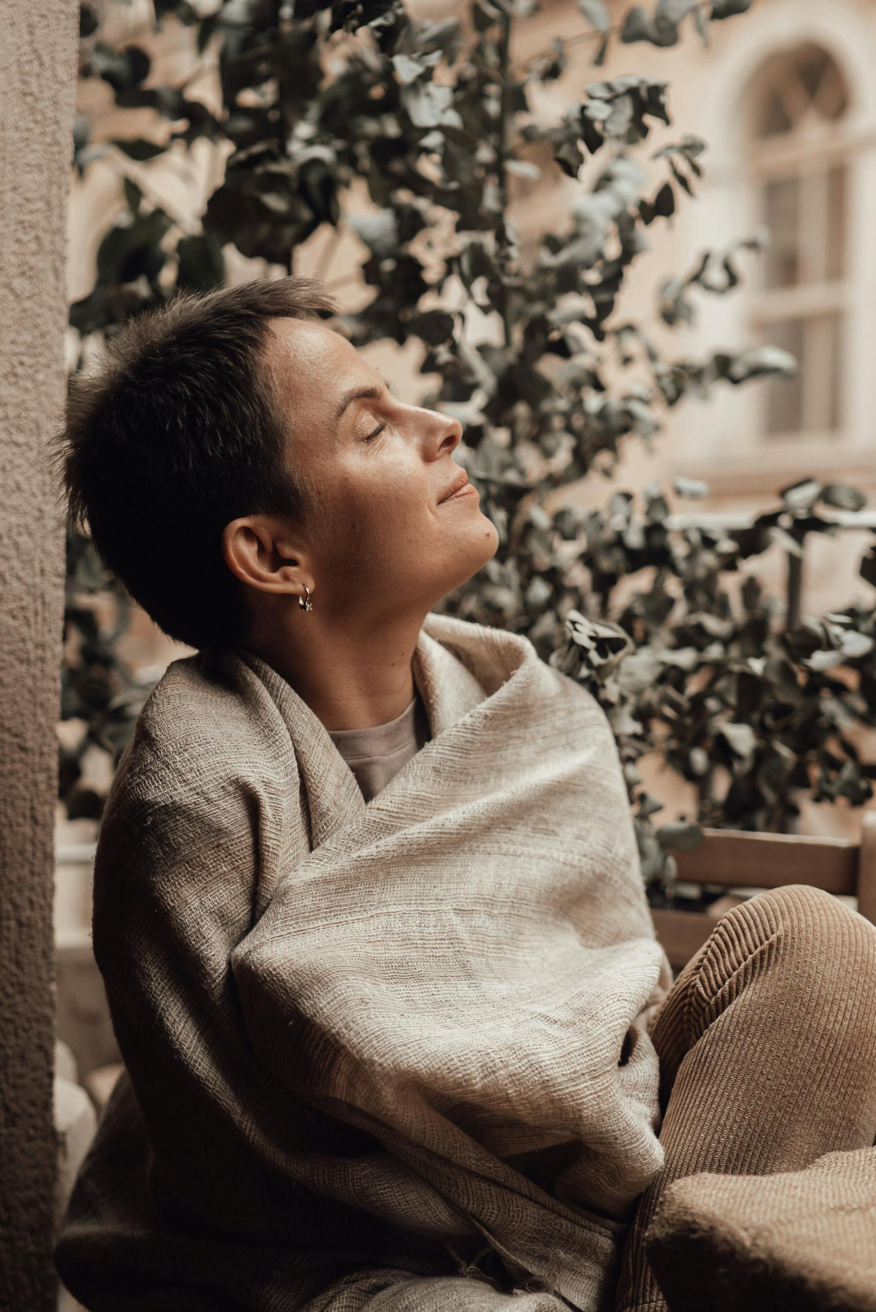 A woman enjoys a peaceful moment on the balcony, wrapped in a cozy blanket, with eyes closed.