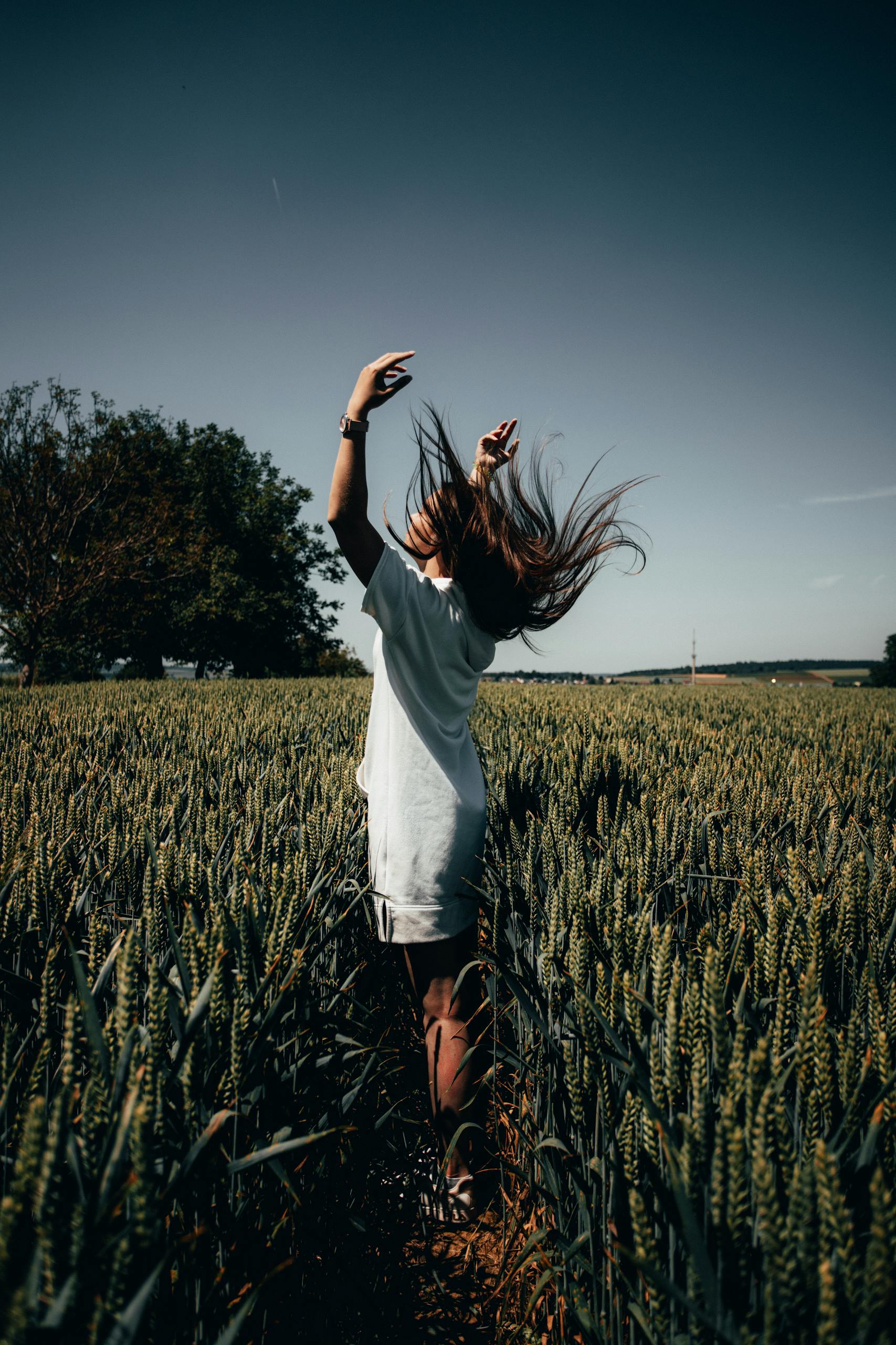 Woman in white dress stands with arms raised in a sunny wheat field, enjoying a bright summer day.